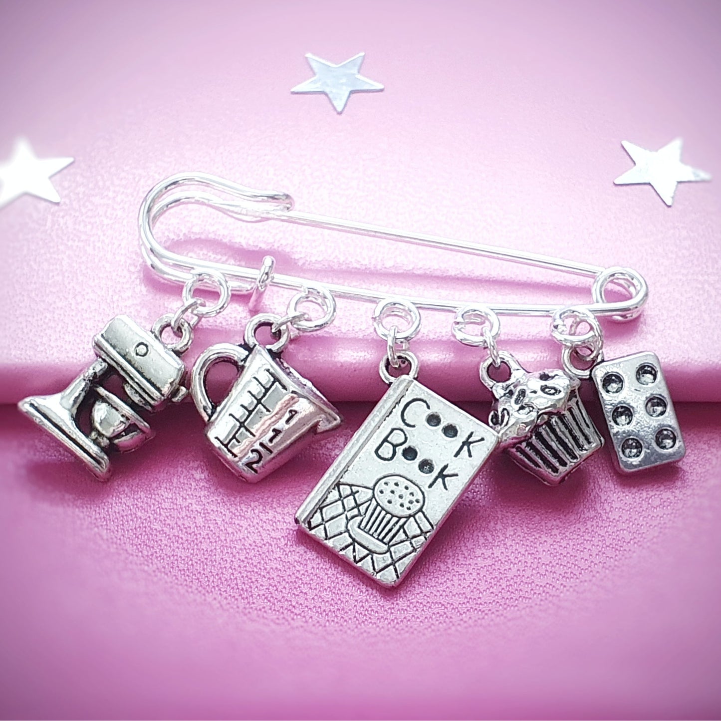 Silver charm brooch with various baking charms on a pink background