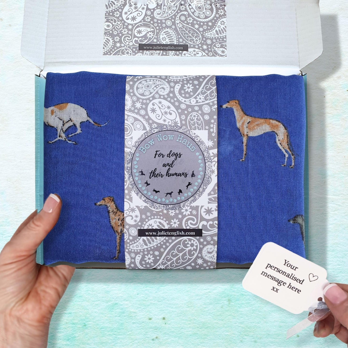 Product packaging with a folded blue fabric scarf featuring dog illustrations inside and a white gift tag with text.