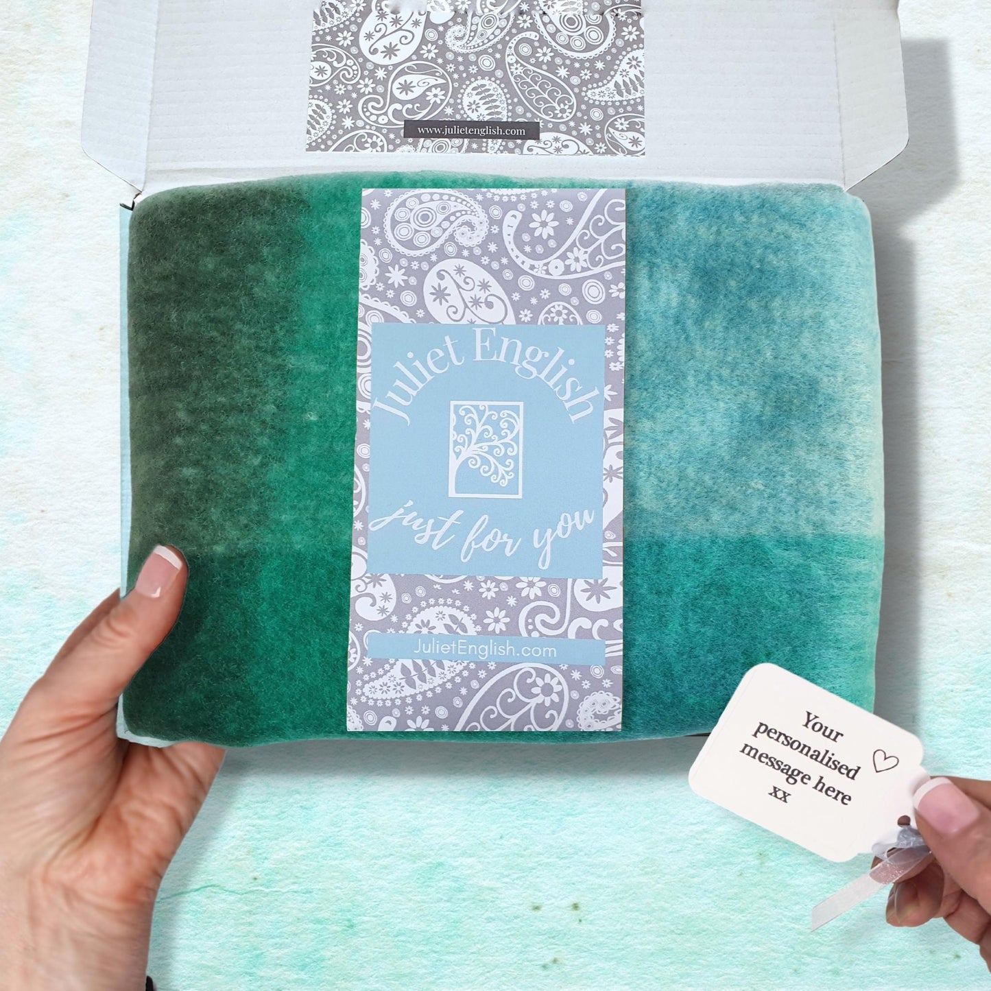 Green check blanket scarf in a box with a label held by a hand on a light blue background