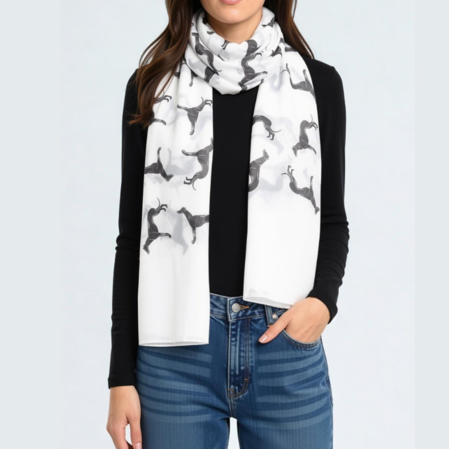 A woman wearing a white scarf with a black greyhound print, styled over a black long-sleeve top and blue jeans against a light gray background.