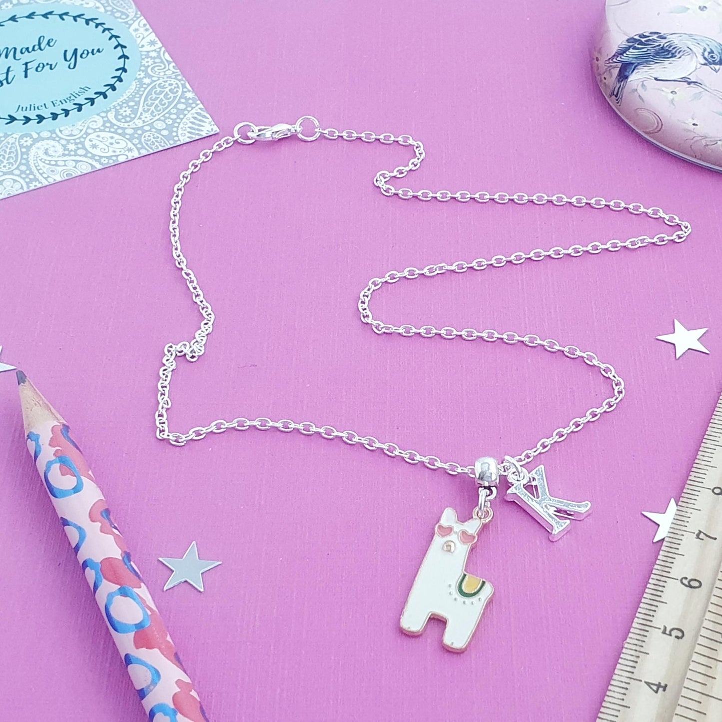 Necklace with llama charm and 'L' pendant on a pink background with stationery items.
