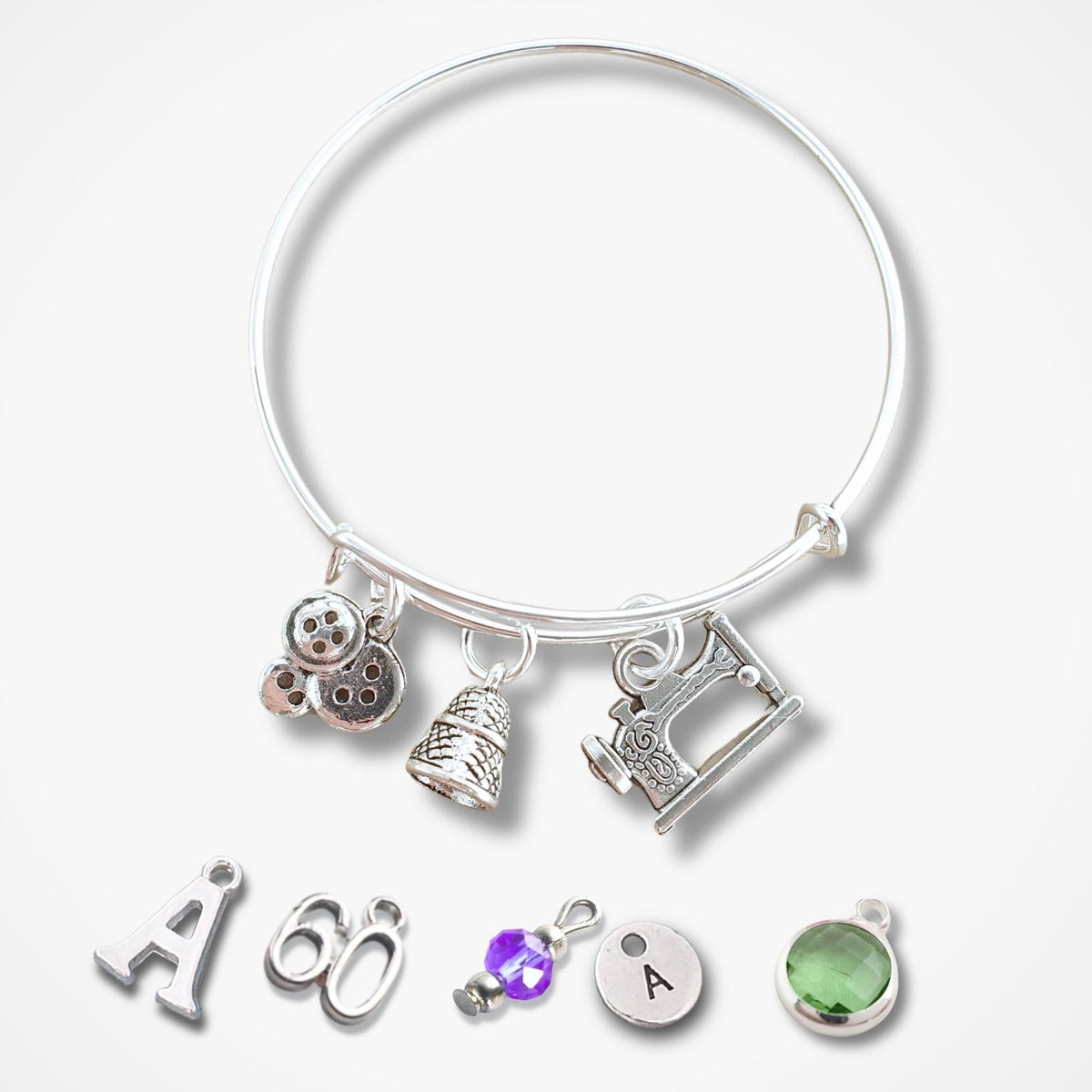 A silver bangle bracelet with multiple charms, including buttons, thimble and sewing machine, and letters/numbers, displayed on a light gray background with additional loose charms below.