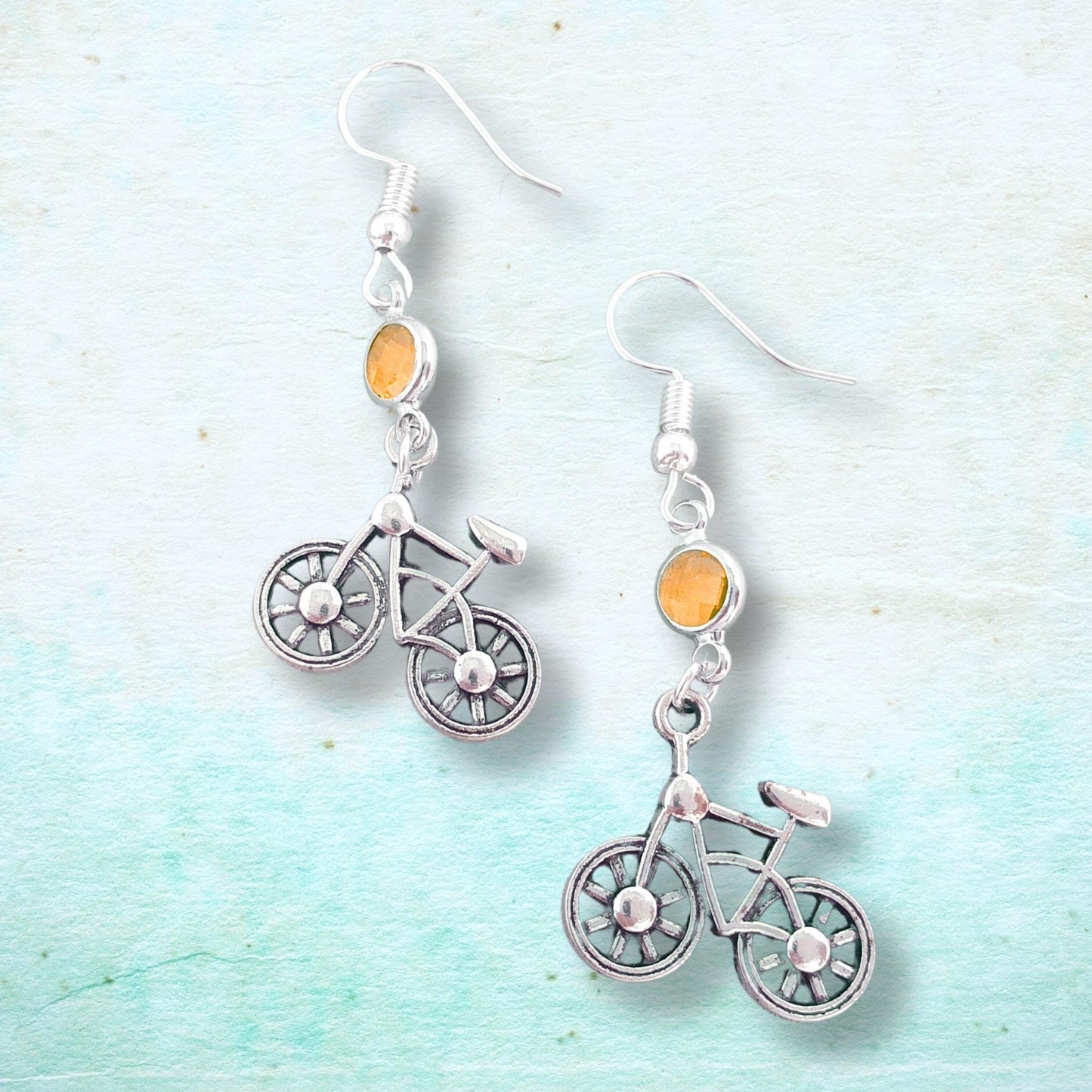 Silver bicycle-shaped earrings with orange beads on a light blue textured background