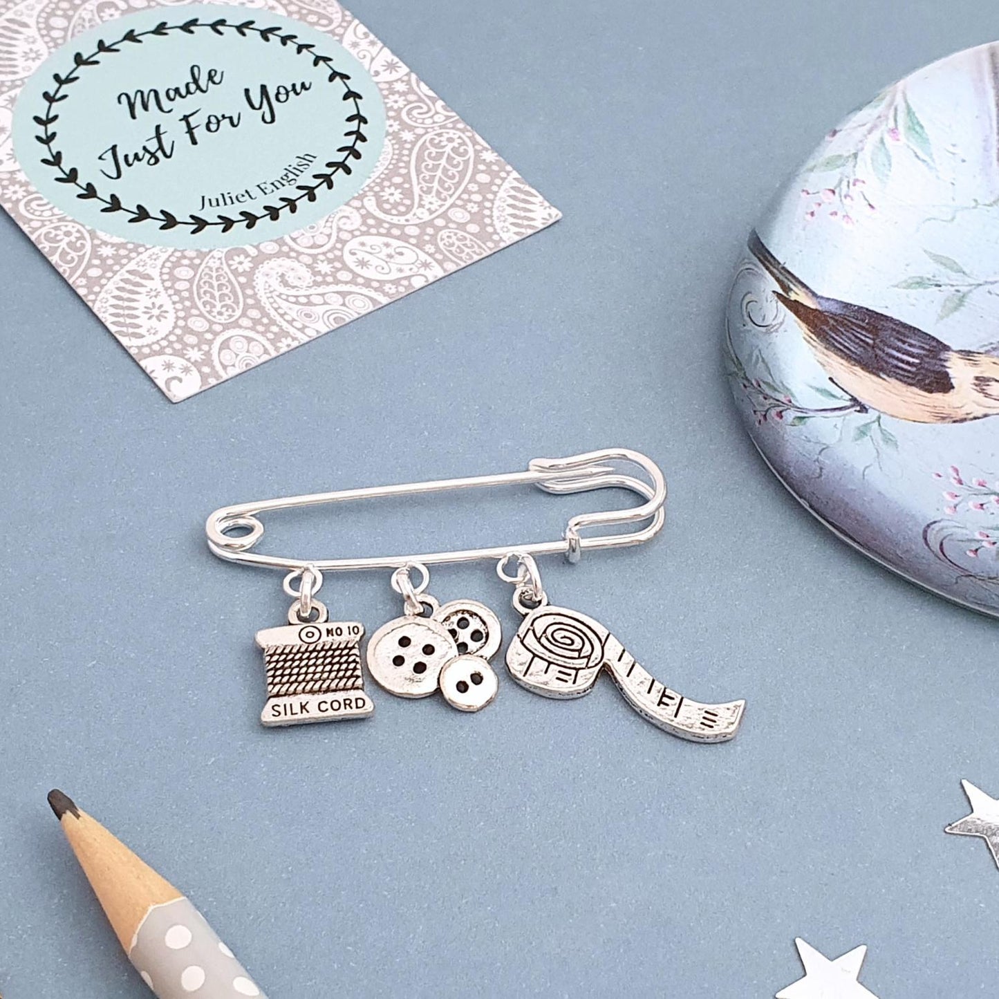 Silver charm brooch with various sewing theme charms on a blue surface with decorative elements