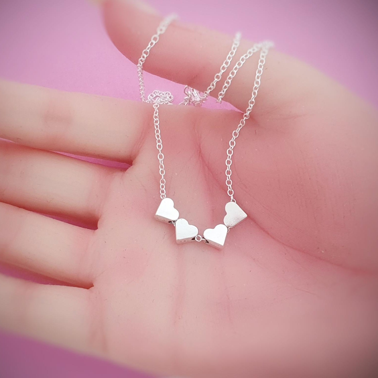 Silver necklace with heart pendants held in a hand against a pink background