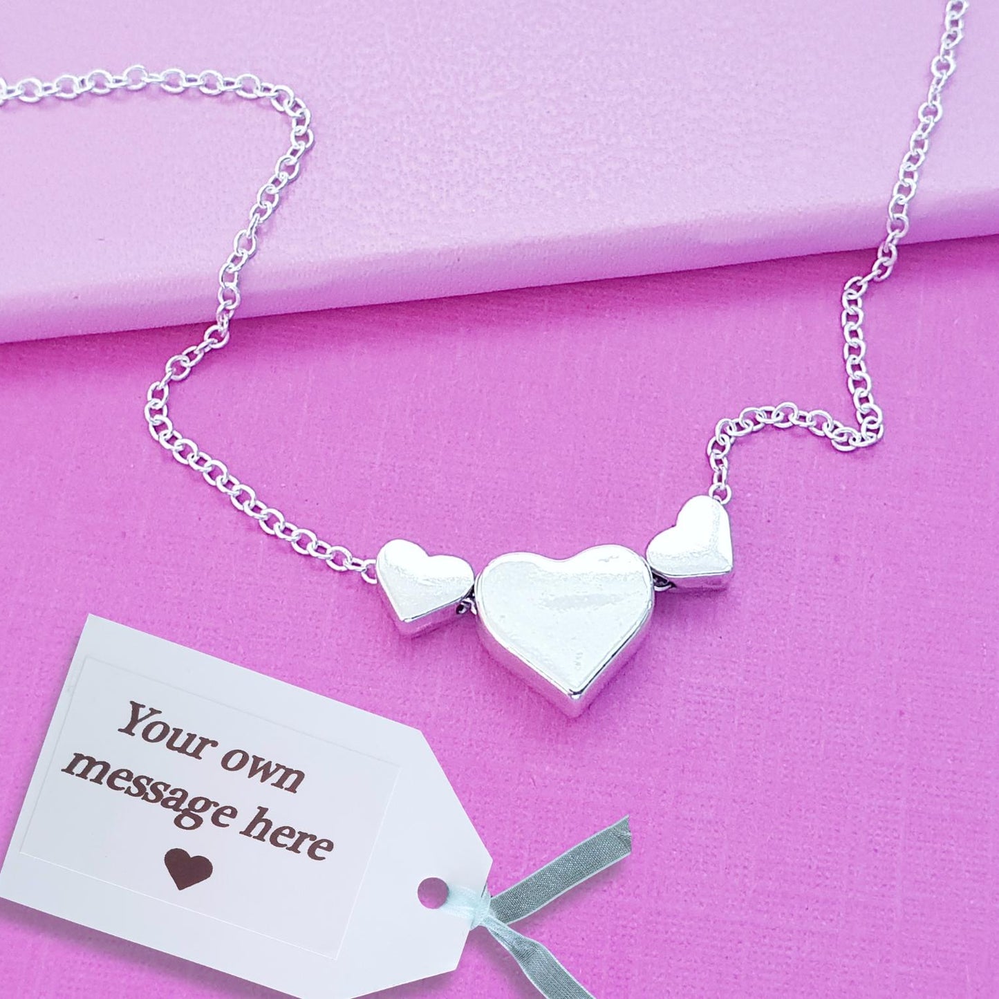 A silver heart necklace with three heart charms, including a larger central heart, displayed on a pink background with a white gift tag that reads 'Your own message here'.