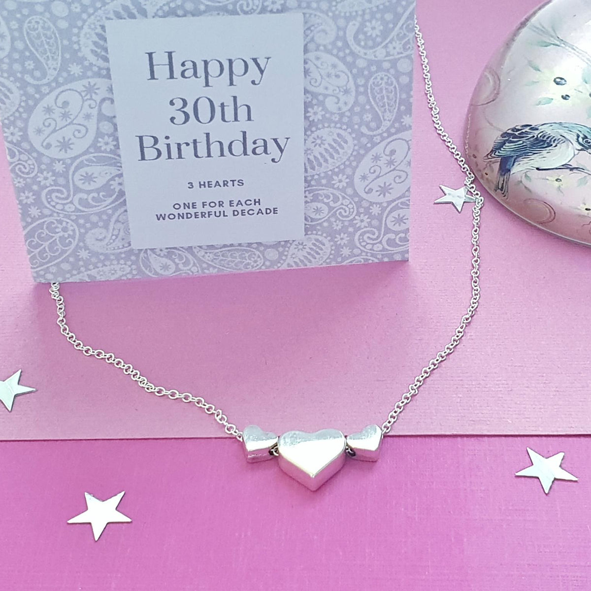 A silver necklace with three interlocking heart charms is displayed on a pink surface next to a grey 'Happy 30th Birthday' card.