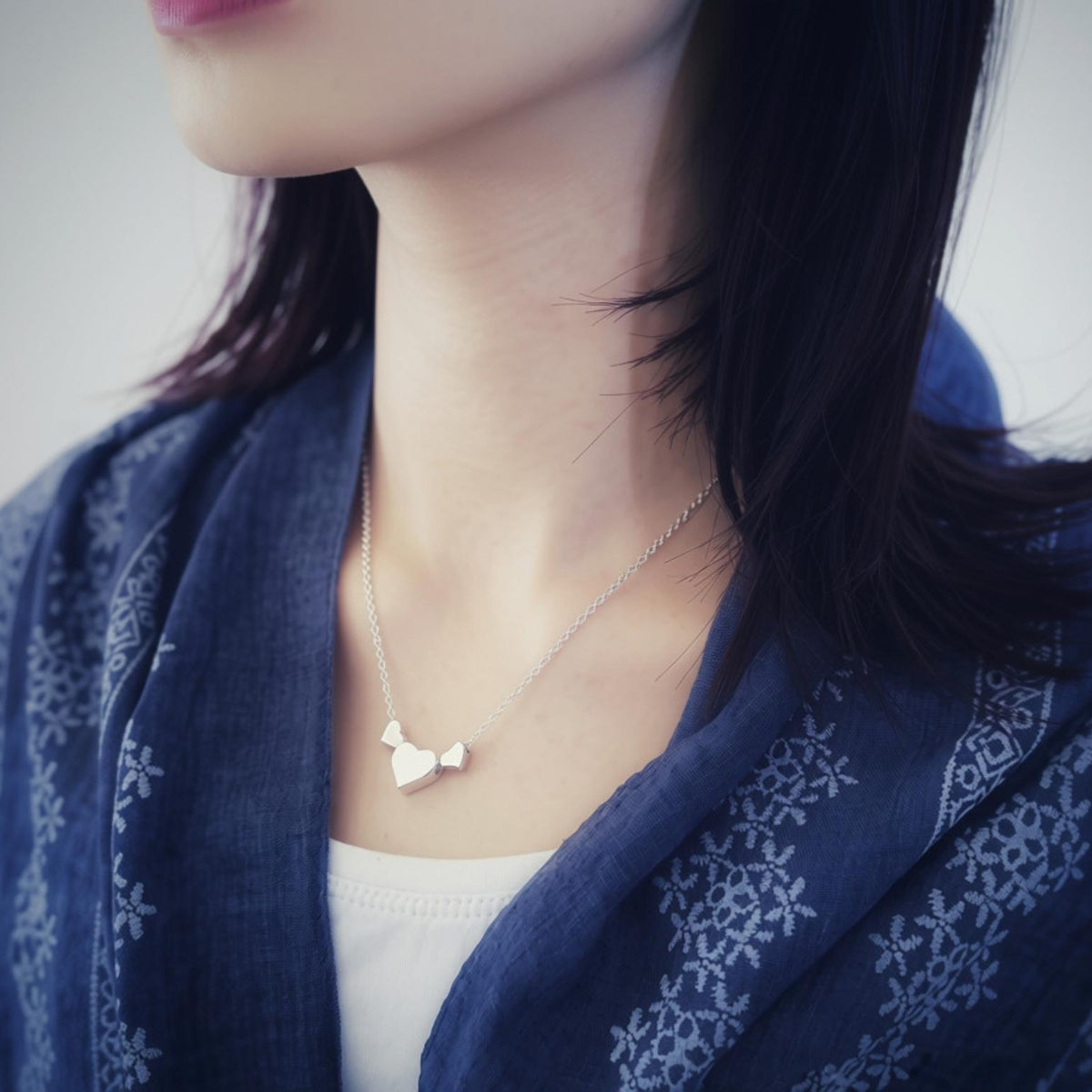 A close-up lifestyle shot of a woman wearing a delicate silver necklace with a small hearts pendant, styled with a dark blue patterned shawl.