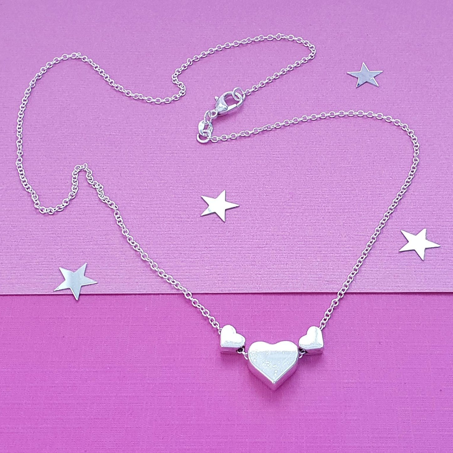 A silver heart necklace with a central heart pendant flanked by two smaller heart charms, displayed on a textured pink background with scattered star-shaped accents.