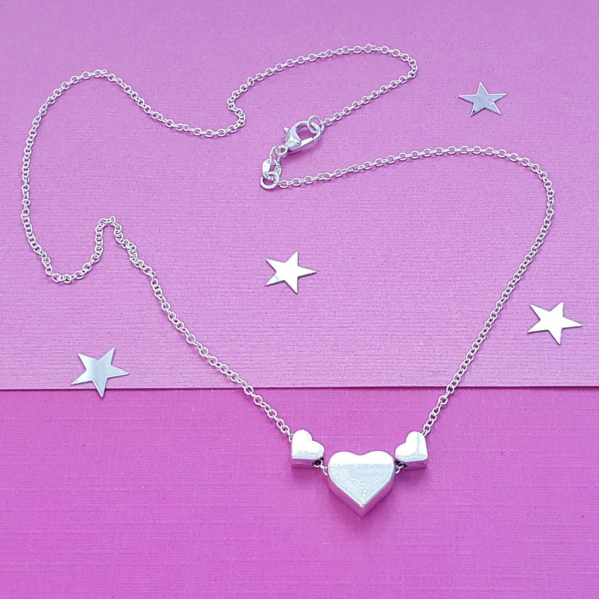 A silver heart necklace with a central heart pendant flanked by two smaller heart charms, displayed on a textured pink background with scattered star-shaped accents.