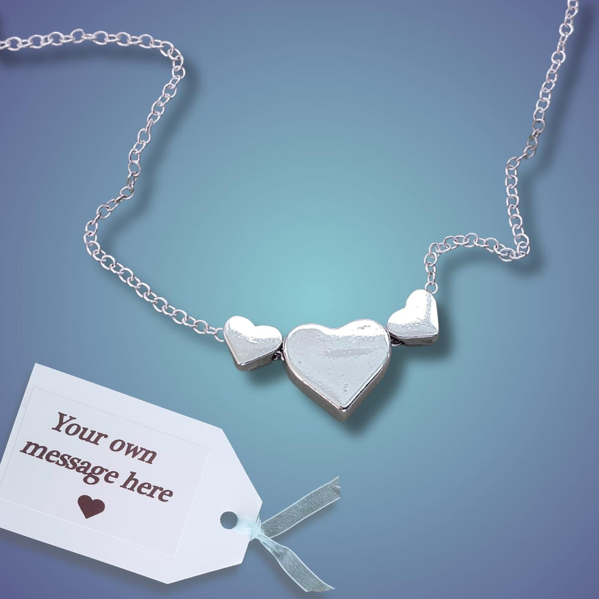 A silver heart necklace with three heart charms on a blue gradient background, shown with a white customizable gift tag.