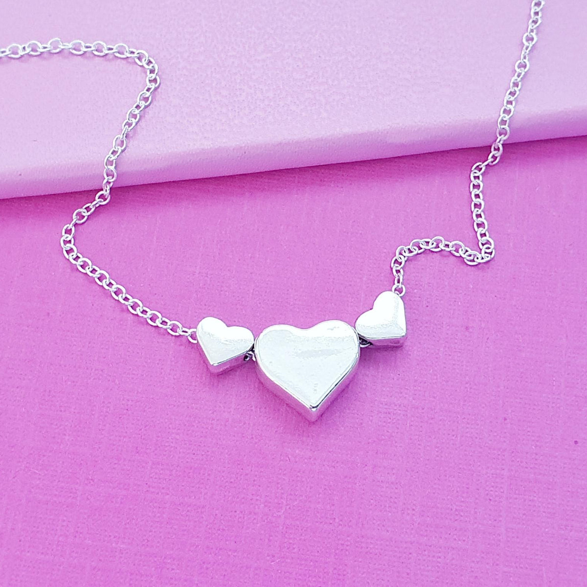 A silver heart necklace with three polished heart charms of varying sizes on a delicate link chain, displayed on a textured pink surface.