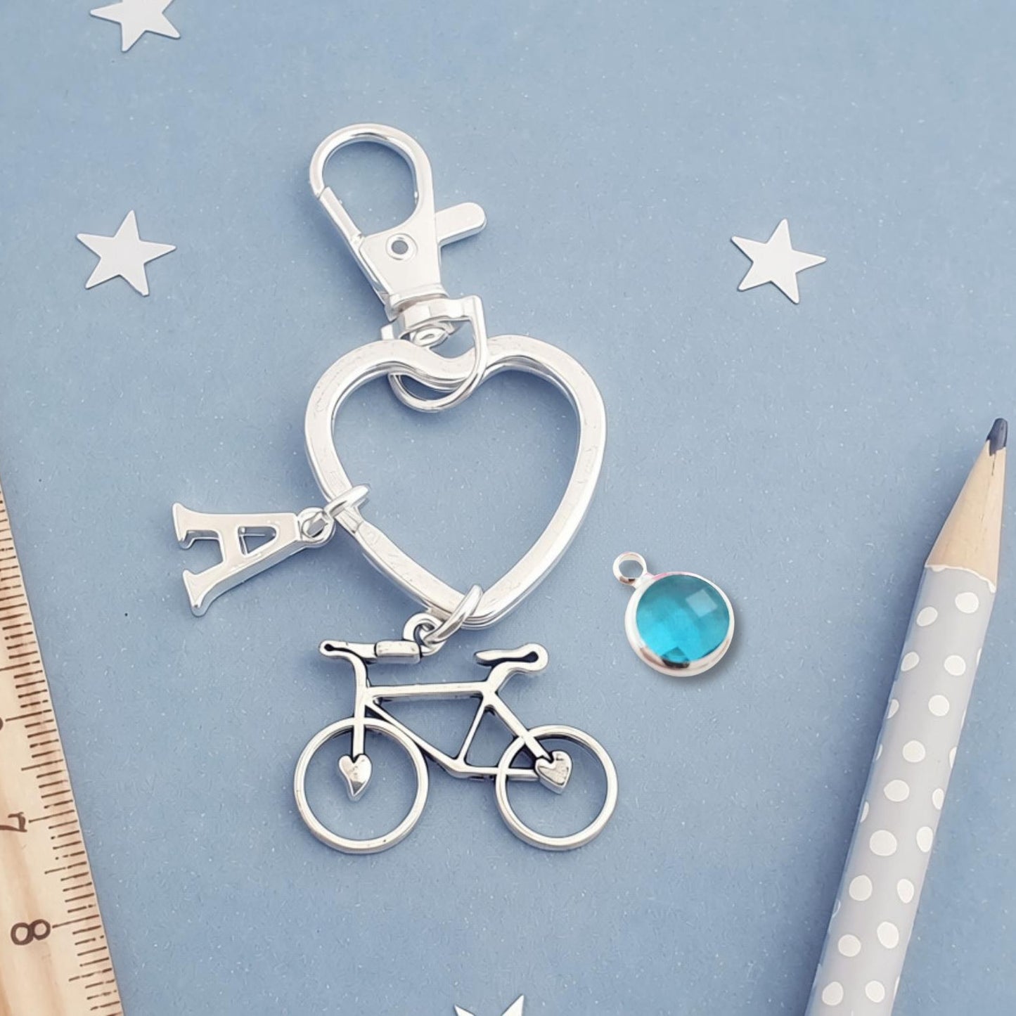 A silver-toned keychain featuring a heart-shaped ring, a bicycle charm, and a letter 'A' charm, displayed on a light blue background with a wooden ruler, star cutouts, and a pencil.