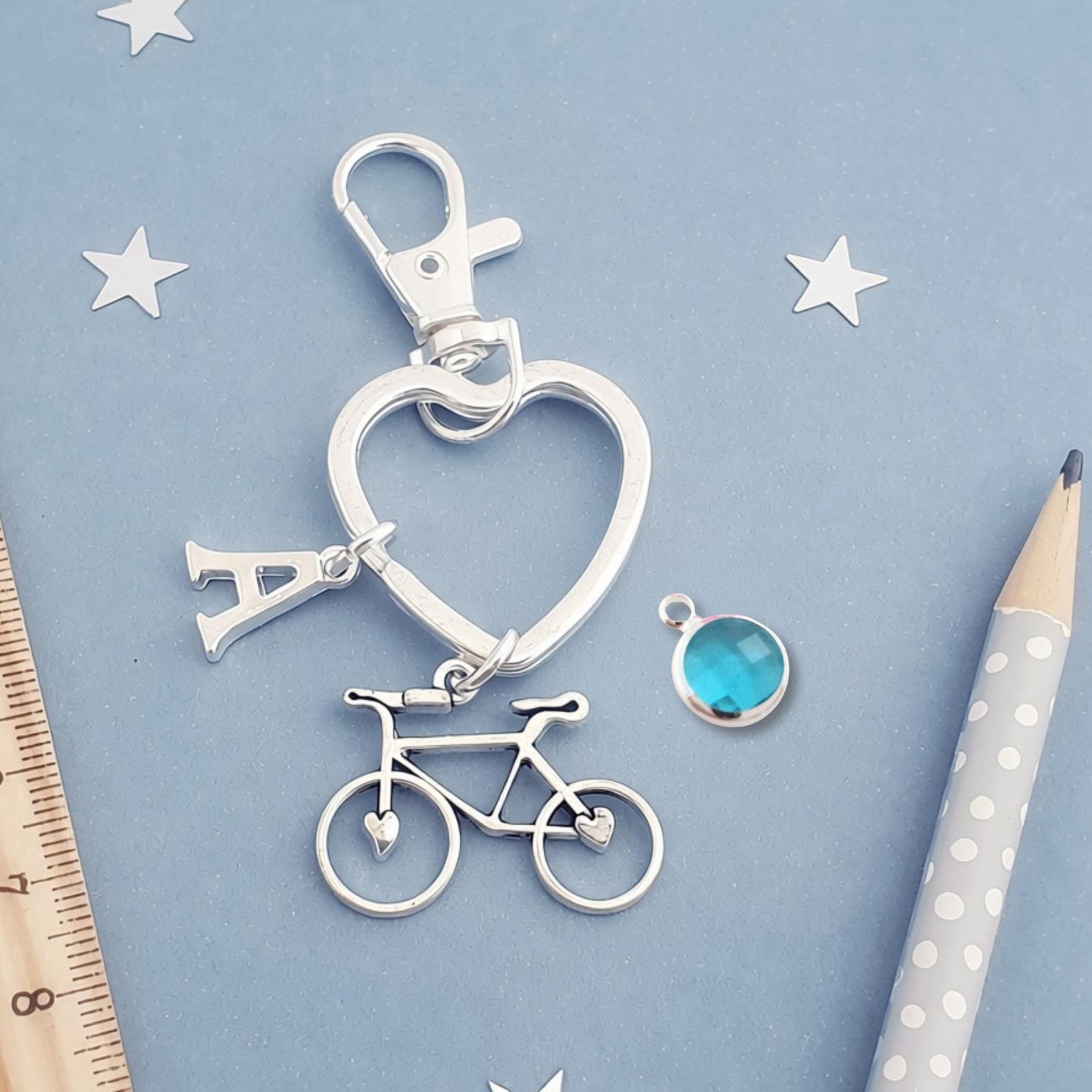 A silver-toned keychain featuring a heart-shaped ring, a bicycle charm, and a letter 'A' charm, displayed on a light blue background with a wooden ruler, star cutouts, and a pencil.