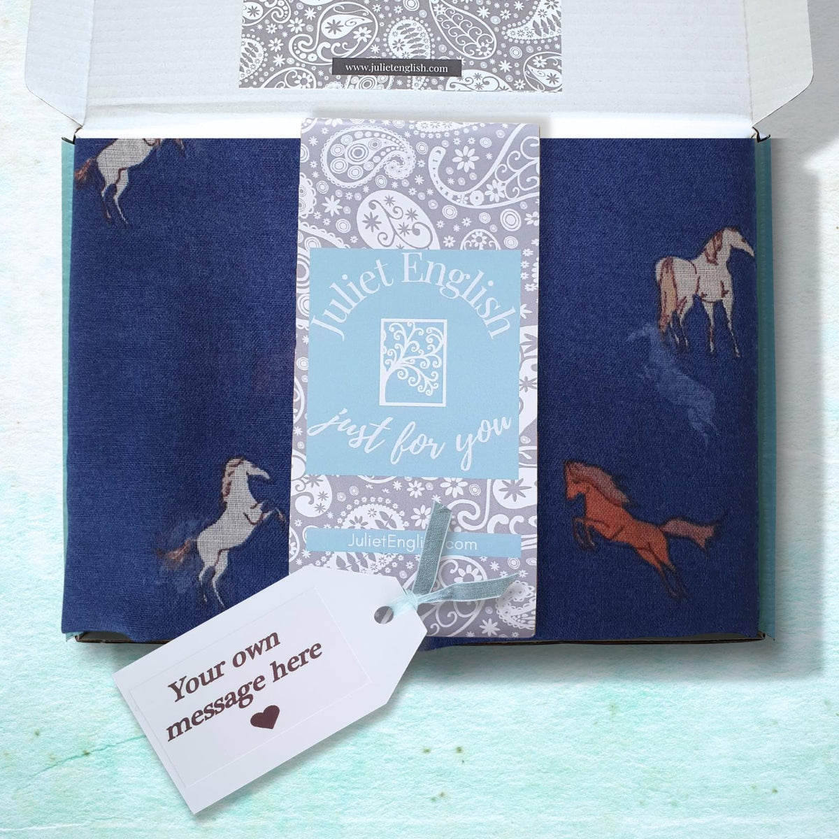 A gift box containing a navy blue folded scarf with horse illustrations, a light blue central card reading 'Juliet English just for you,' and a white gift tag that says 'Your own message here,' shown on a pale aqua textured background.