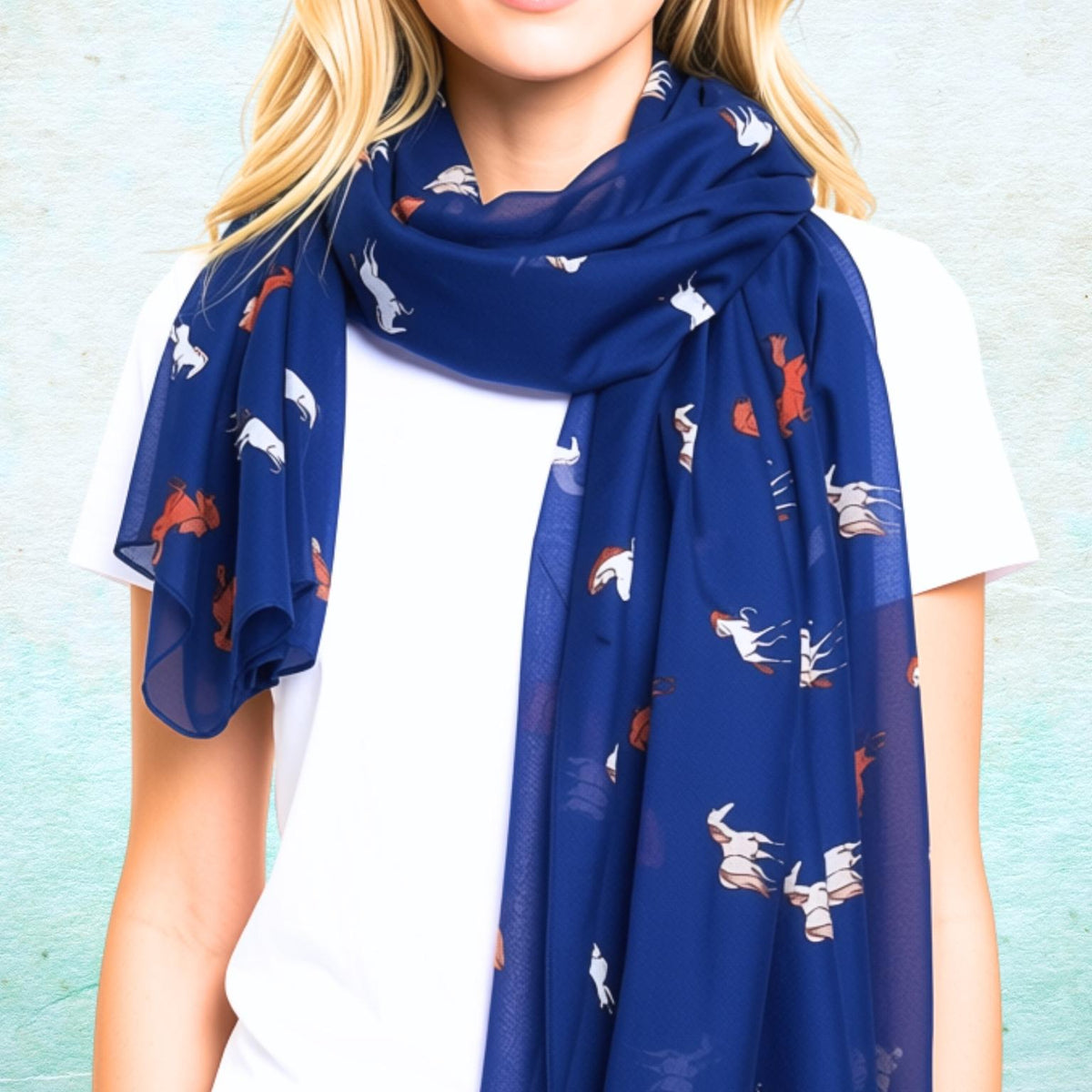 A woman wearing a navy blue scarf with a pattern of white and orange horses, styled over a white t-shirt against a textured light blue wall.