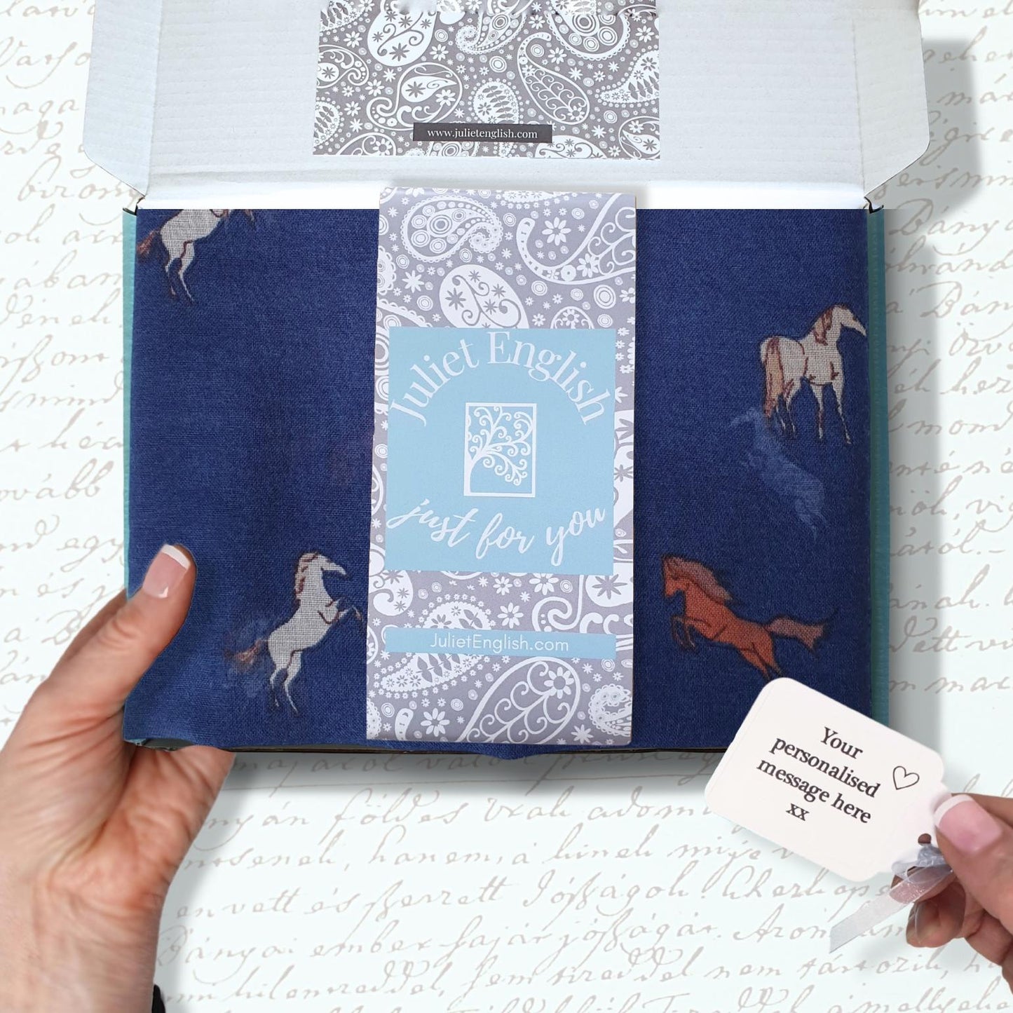 A hand holds an open white gift box containing a navy blue fabric scarf with horse illustrations and a central light blue label reading 'Juliet English just for you.' A small white tag is shown in the foreground against a white background with faint cursive writing.