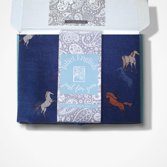 An open gift box containing a navy blue scarf with horse illustrations and a central light blue band that reads 'Juliet English just for you' in white script, surrounded by and a paisley pattern.