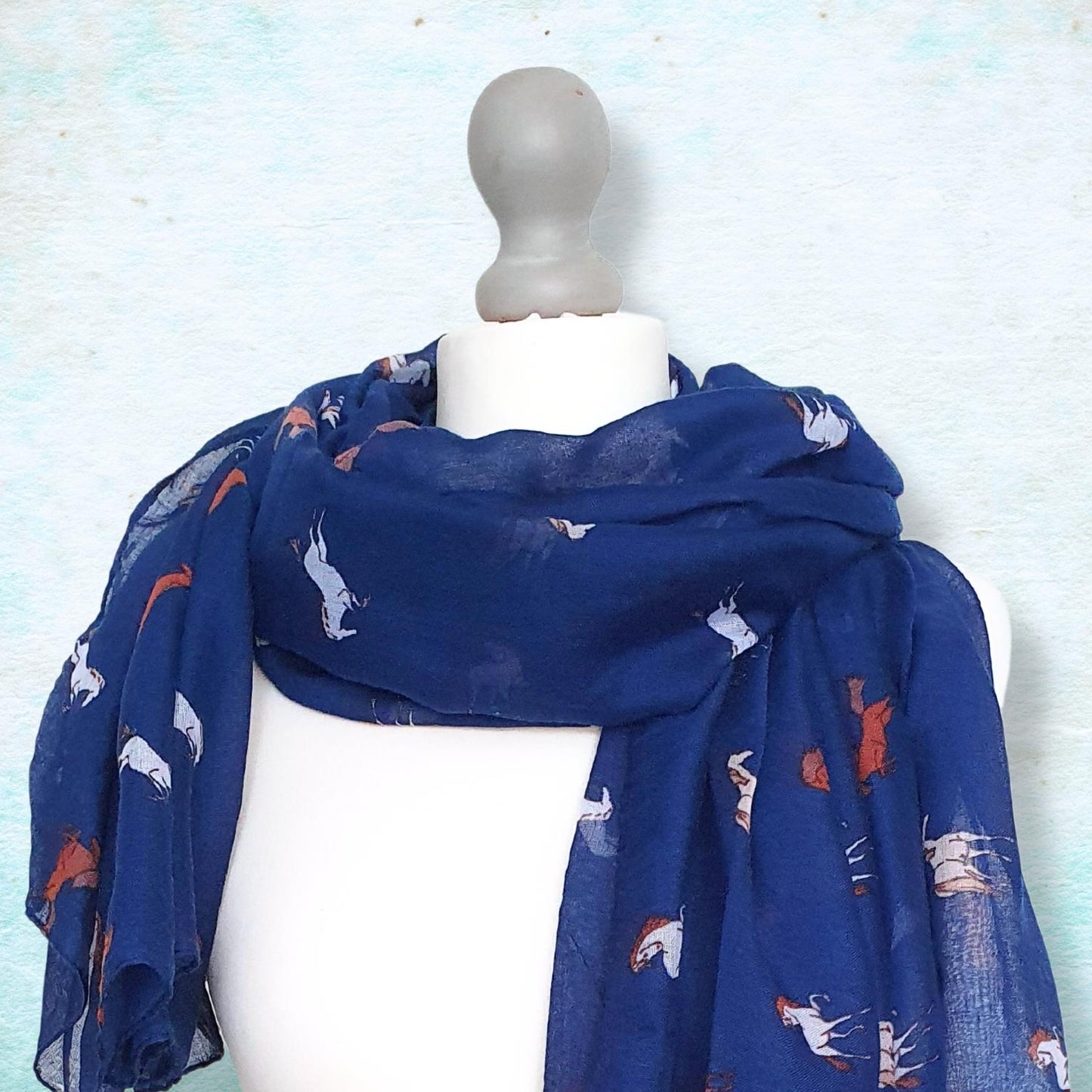 A navy blue scarf with a pattern of white and orange horses, draped on a white mannequin torso against a textured light blue background.