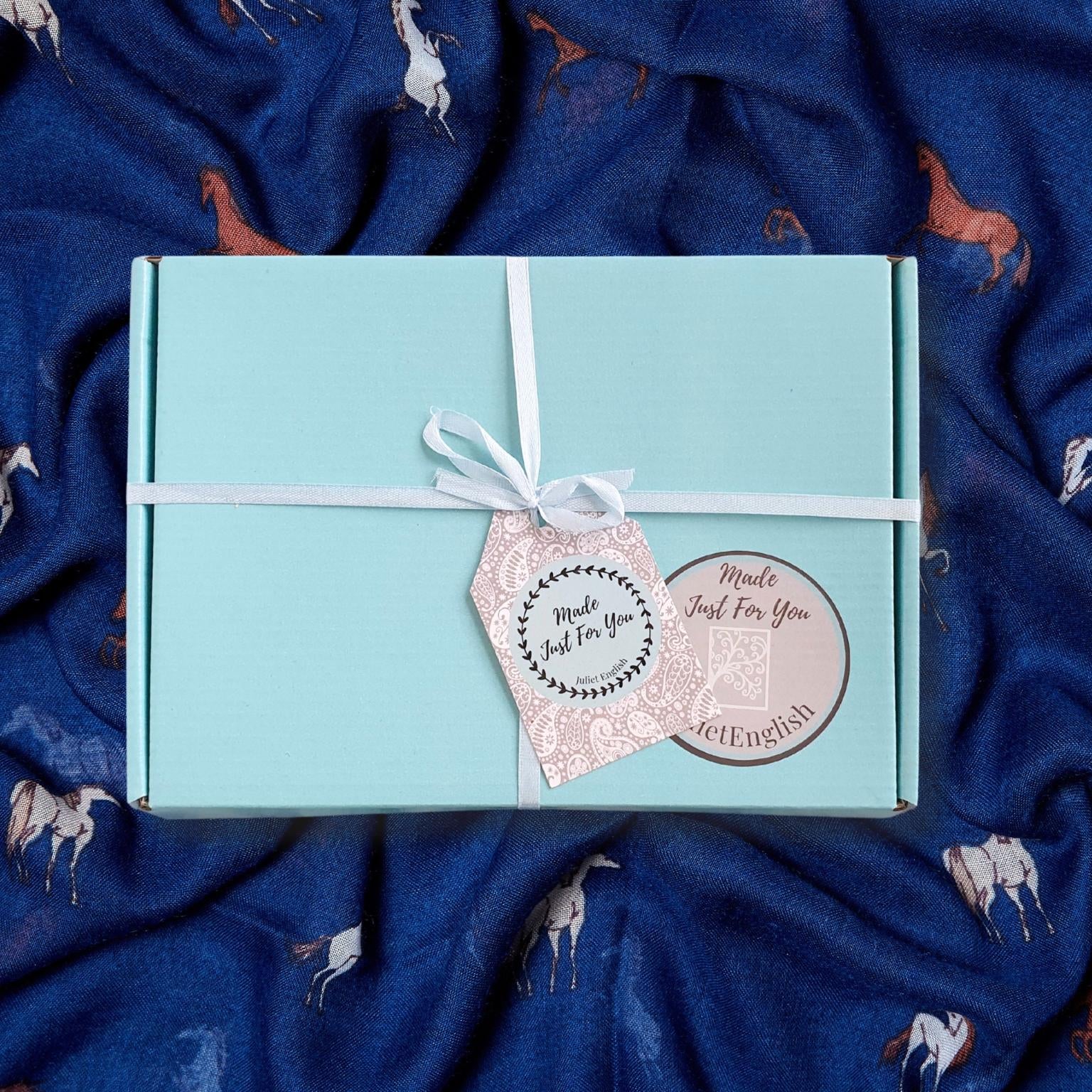 A light blue gift box tied with a white ribbon and decorated with two decorative tags, placed on a navy blue scarf background with a horse pattern.