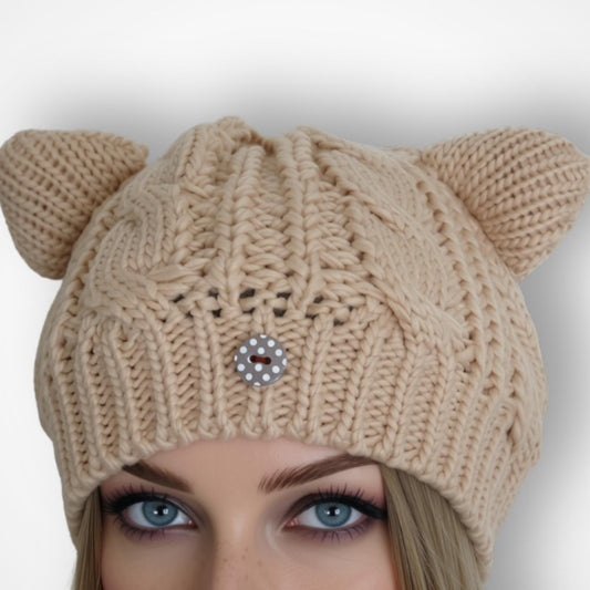 A beige cable-knit beanie hat with two small cat ears on top and a decorative dotty button at the center front, shown on a model against a light gray background.