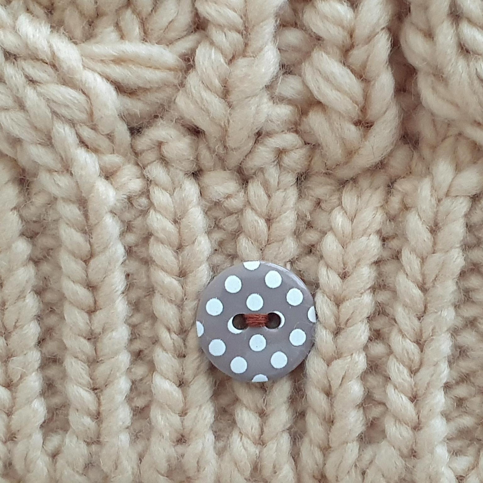 Close-up of a chunky cream-colored cable-knit fabric with a single gray button featuring white polka dots.