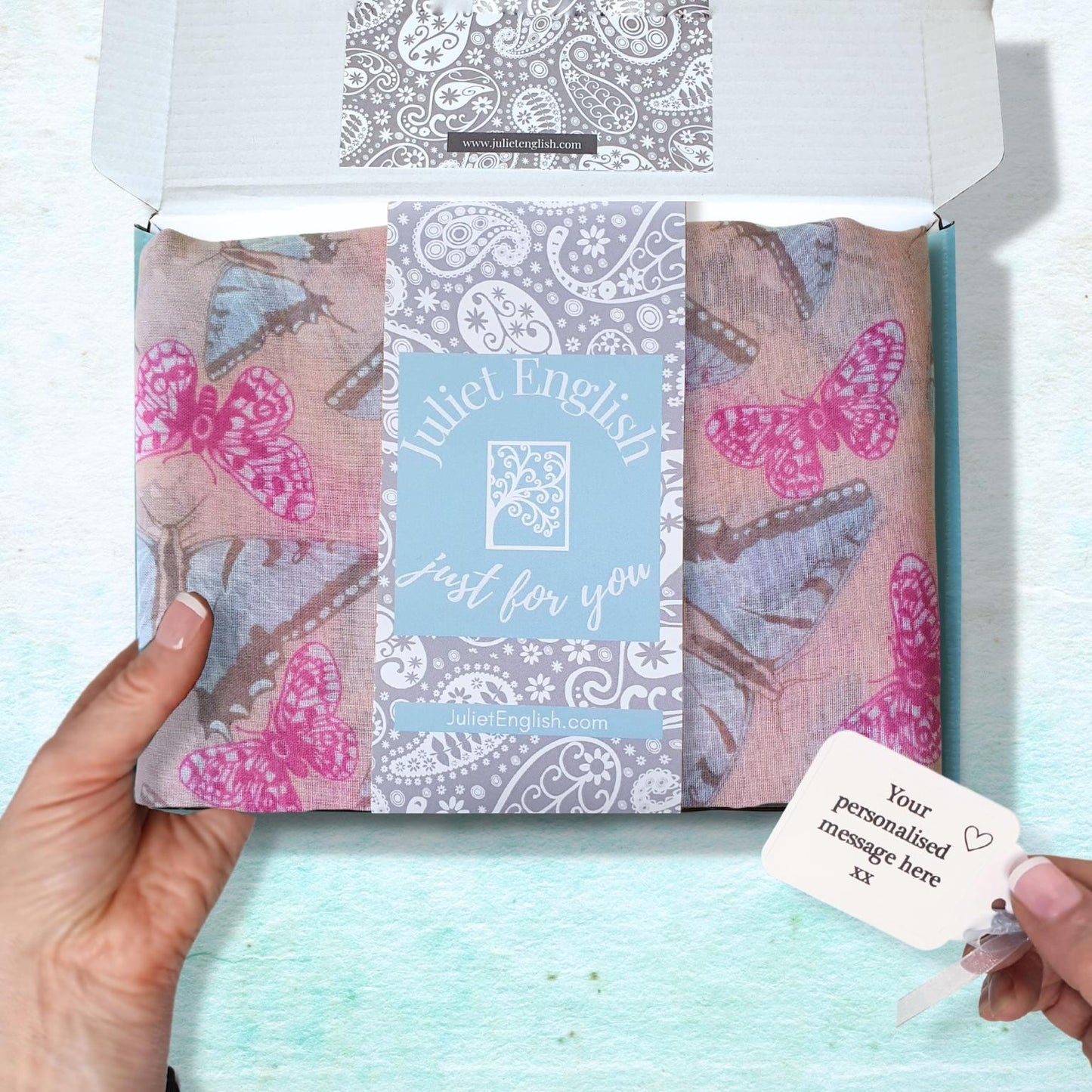 A hand holds an open white gift box containing a folded patterned scarf with pink and blue butterflies, a central blue card reading 'Juliet English just for you', and a small personalized gift tag held by another hand, set against a light aqua textured background.