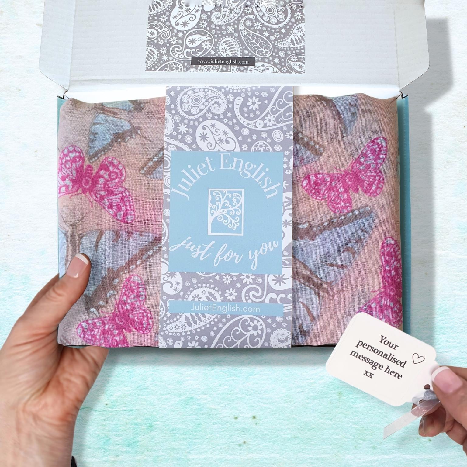 A hand holds an open white gift box containing a folded patterned scarf with pink and blue butterflies, a central blue card reading 'Juliet English just for you', and a small personalized gift tag held by another hand, set against a light aqua textured background.