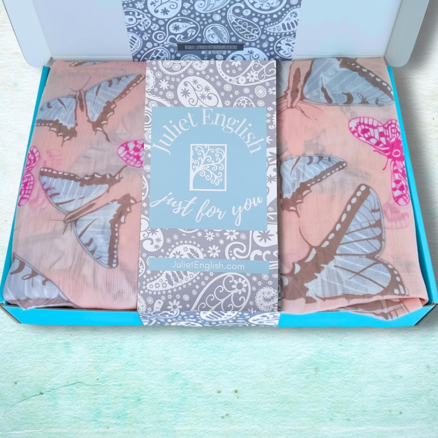 An open turquoise gift box containing a folded pink butterfly-patterned scarf with a central light blue card that reads 'Juliet English just for you'.