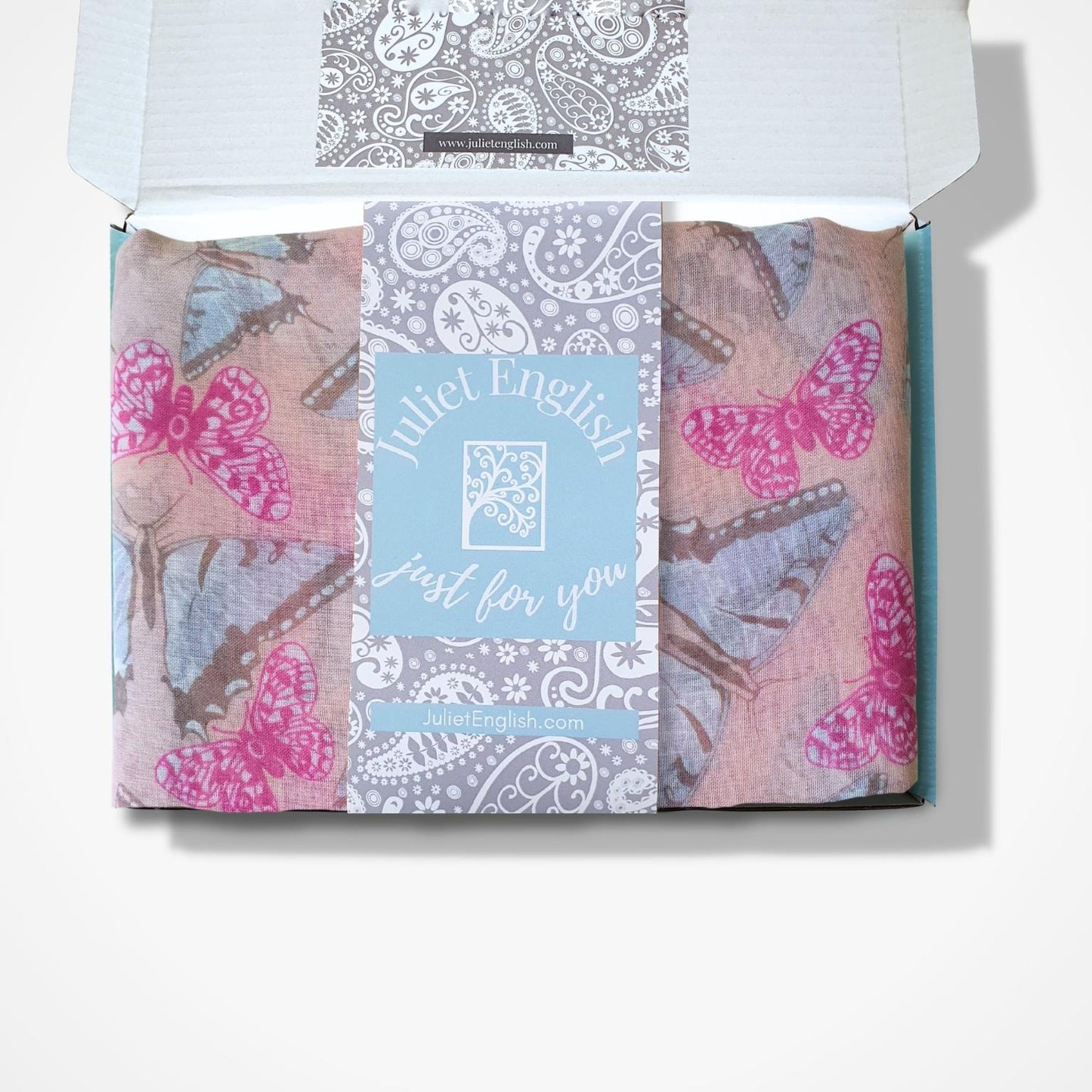 A folded butterfly patterned scarf in an open white box, featuring pink and blue butterfly prints on a dusty pink background with a central light blue card reading 'Juliet English Just for you'.