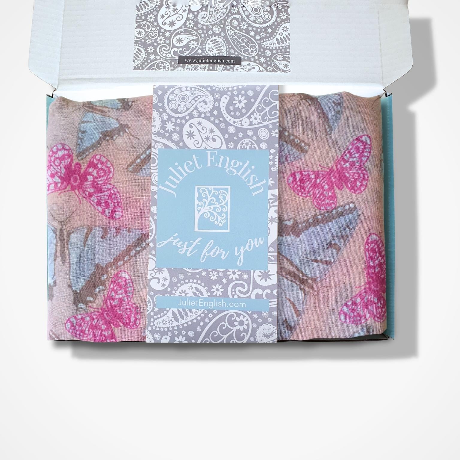 A folded butterfly patterned scarf in an open white box, featuring pink and blue butterfly prints on a dusty pink background with a central light blue card reading 'Juliet English Just for you'.