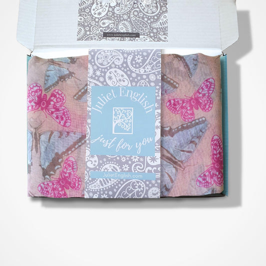 A folded butterfly patterned scarf in an open white box, featuring pink and blue butterfly prints on a dusty pink background with a central light blue card reading 'Juliet English Just for you'.