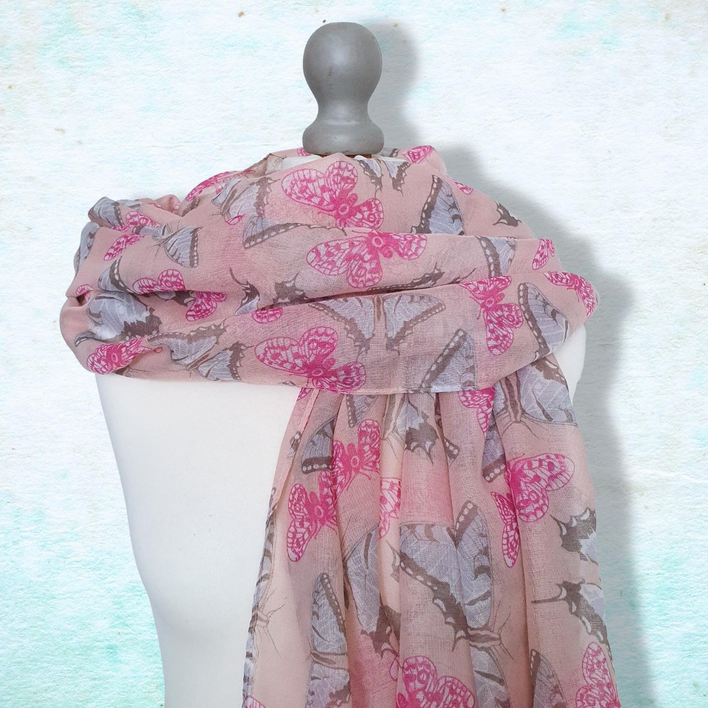 A lightweight pink scarf with a gray and magenta butterfly pattern, draped on a white mannequin torso against a textured light blue background.