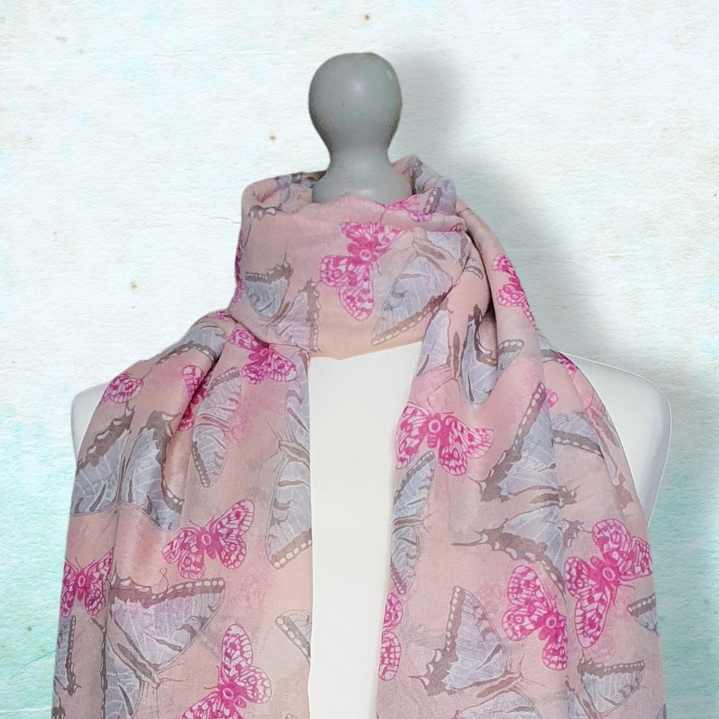 A lightweight pink scarf with a butterfly print draped on a white mannequin torso against a textured light blue background.