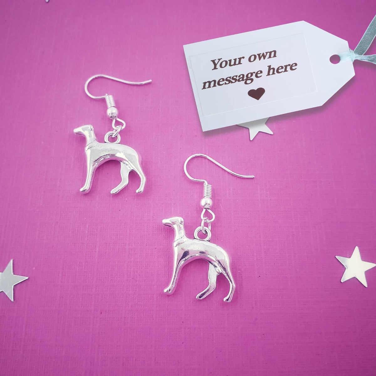 A pair of silver Italian Greyhound dog drop earrings displayed on a textured pink background with decorative silver stars and a white gift tag that reads 'Your own message here'.