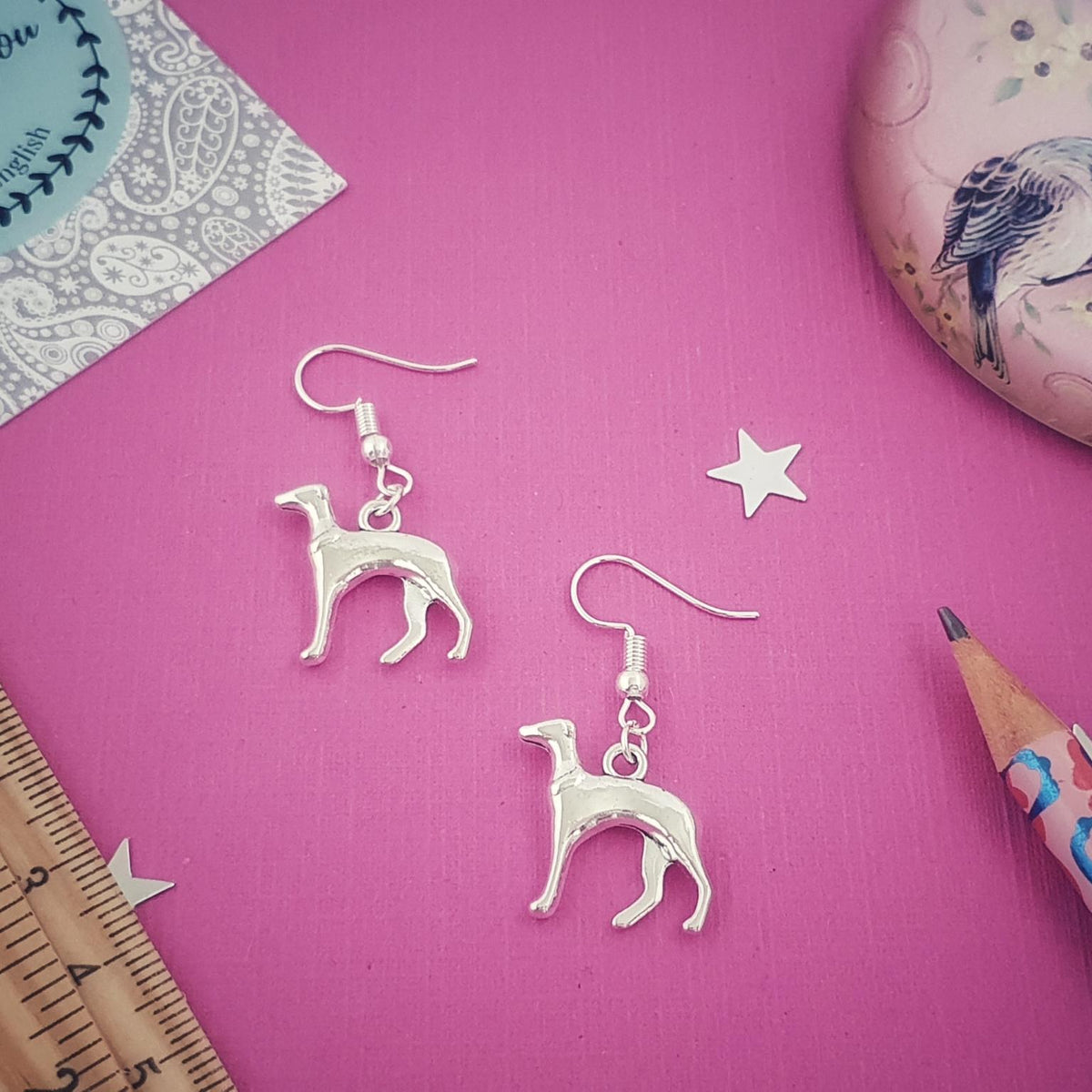A pair of silver-tone dangle earrings shaped like whippet dogs, arranged on a bright pink background with decorative props including a ruler, pencil, star cutouts, and a patterned card.