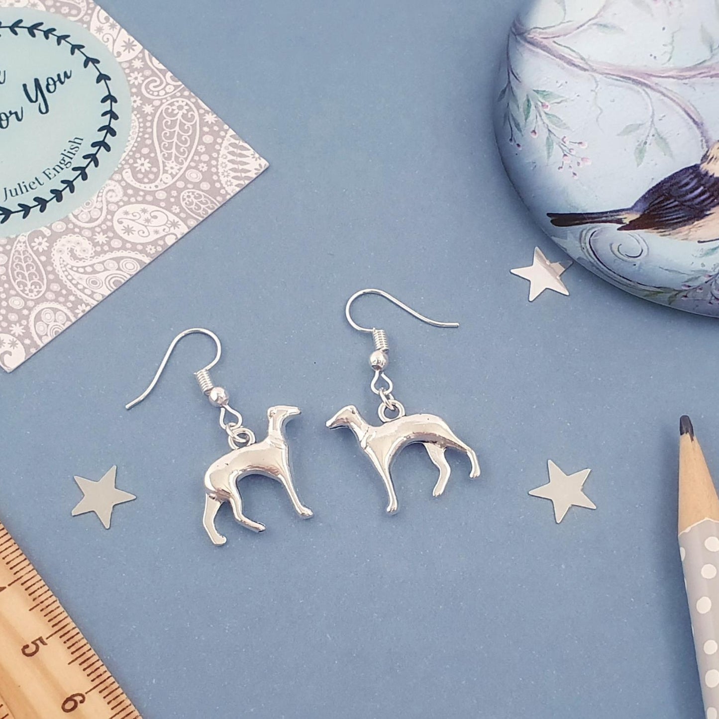 A pair of silver dangling earrings shaped like slender dogs, displayed on a dusty blue background with decorative props including a wooden ruler, a pencil, star cutouts, a patterned card, and a bird-themed tin.