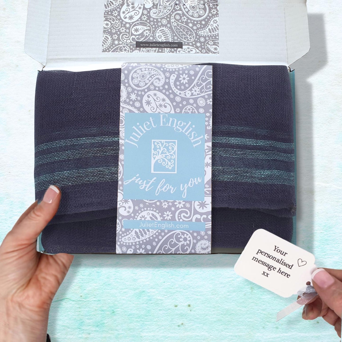 Navy blue striped scarf in a box with a label held by a hand on a light blue background