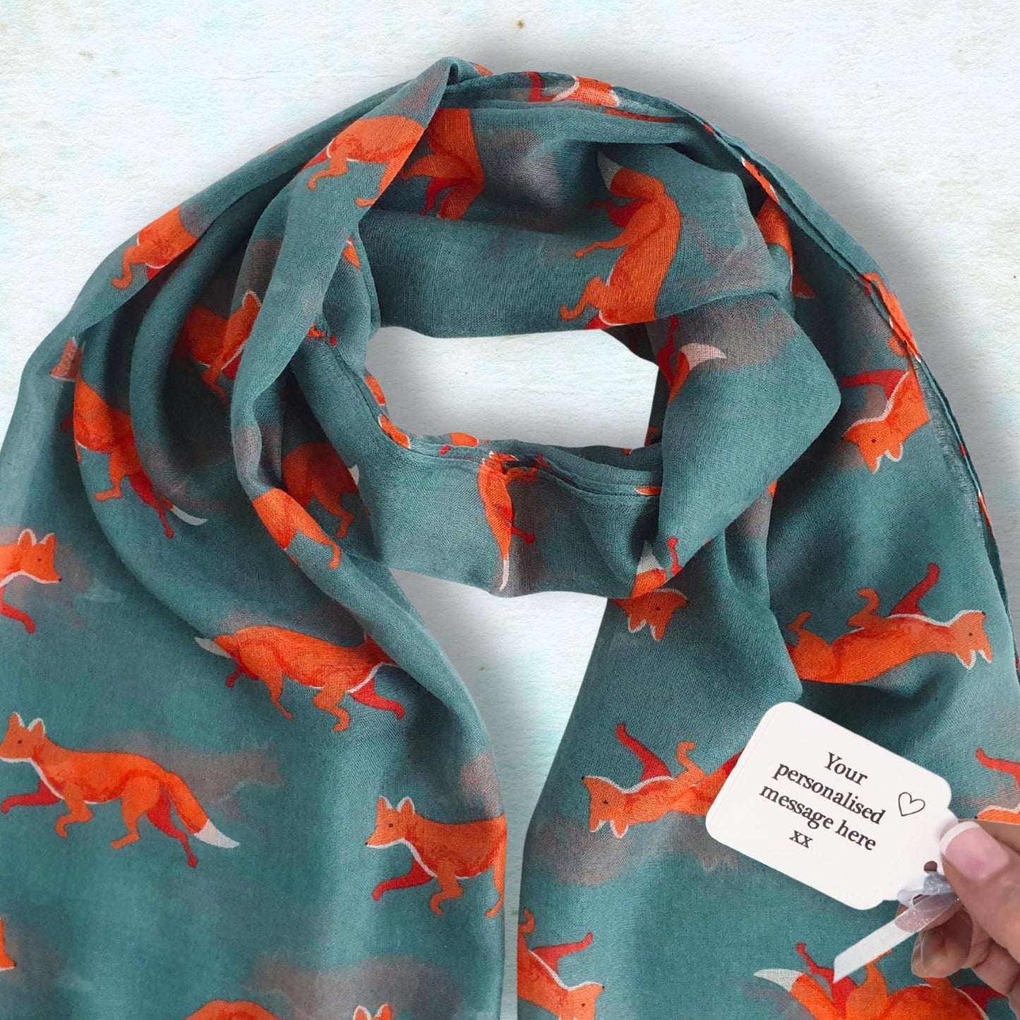 A teal scarf with an all-over orange fox print, draped on a light blue textured background. A hand is holding a white paper tag attached to the scarf.