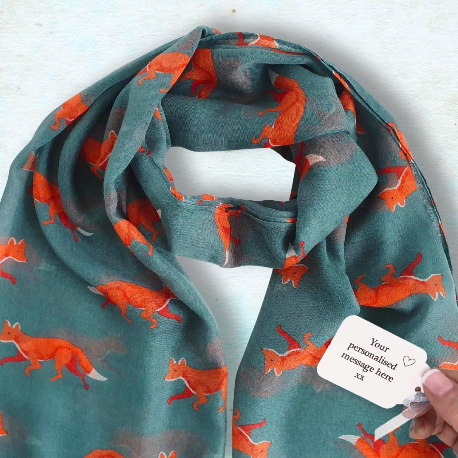 A teal scarf with an all-over orange fox print, draped on a light blue textured background. A hand is holding a white paper tag attached to the scarf.