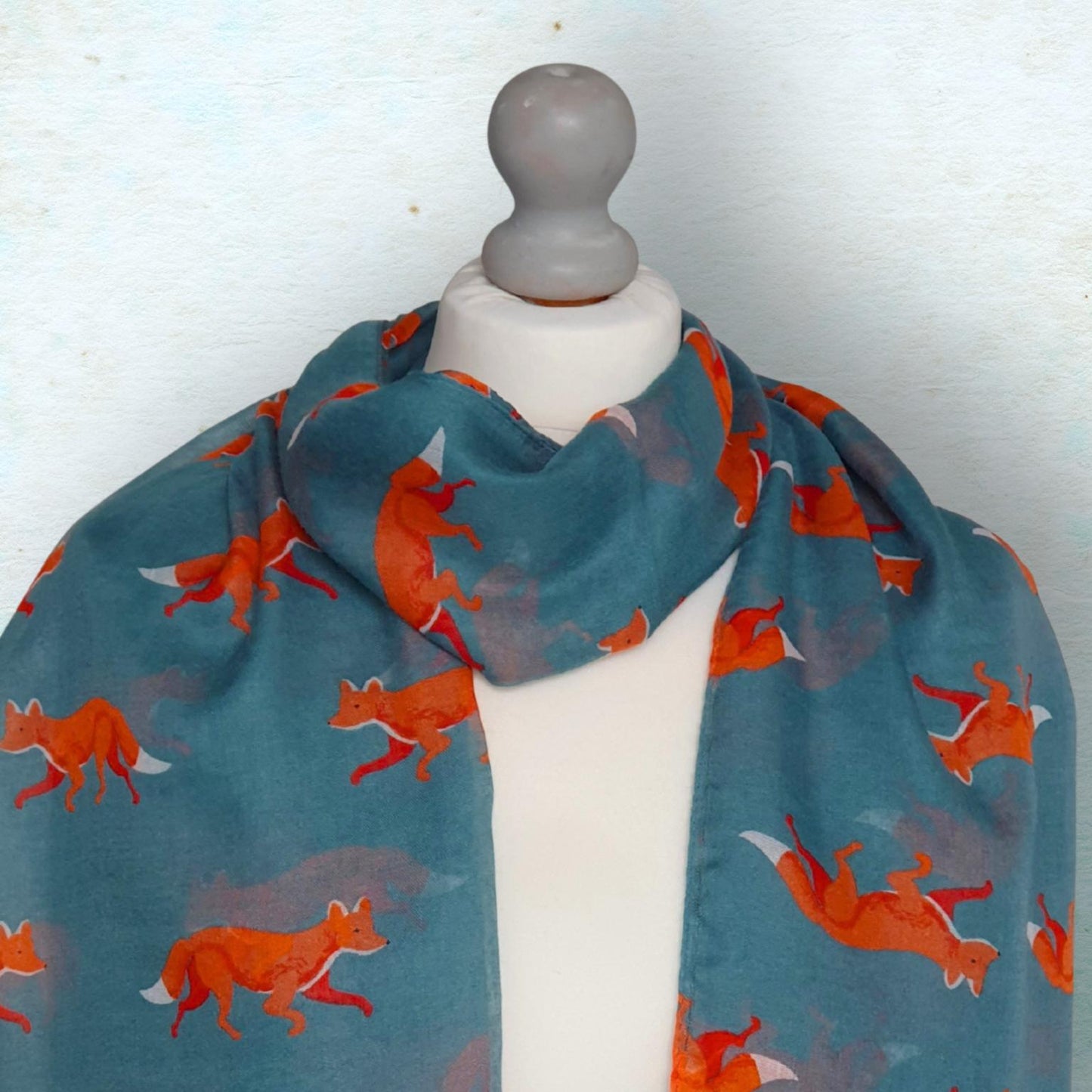 A teal green scarf with a repeating pattern of orange foxes, displayed on a white mannequin torso against a textured light blue background.