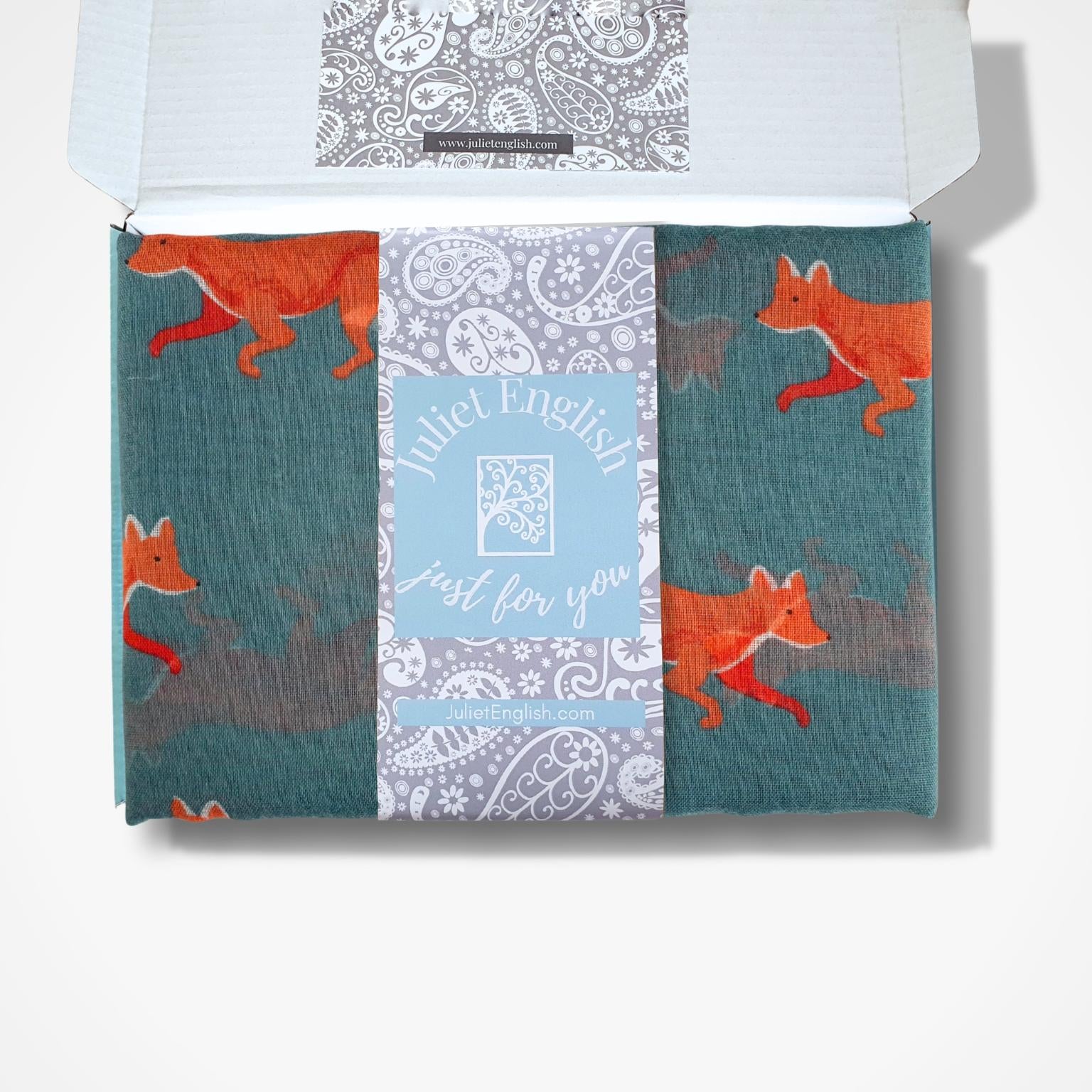 A teal green scarf with a repeating pattern of orange foxes in a box
