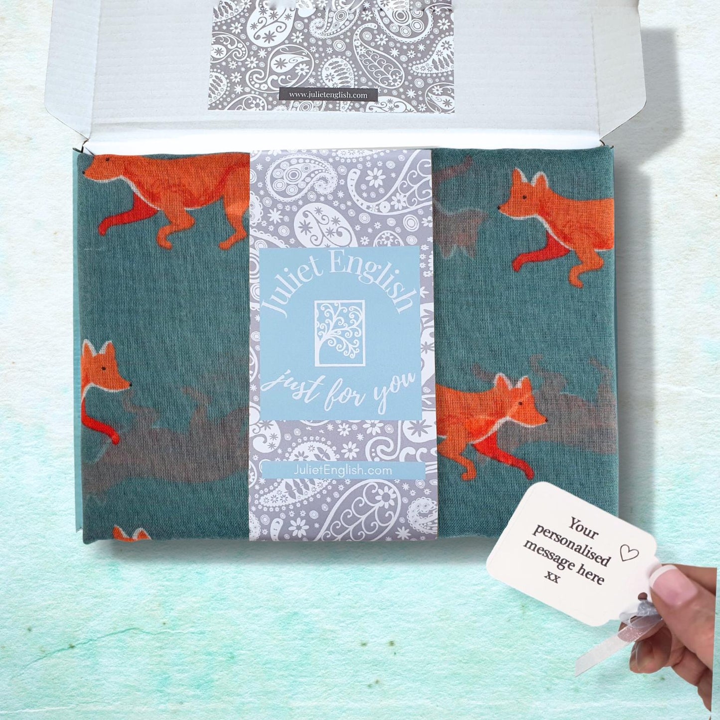A  box on a light aqua textured background containing a teal green scarf with orange foxes and a central decorative band reading 'Juliet English just for you."