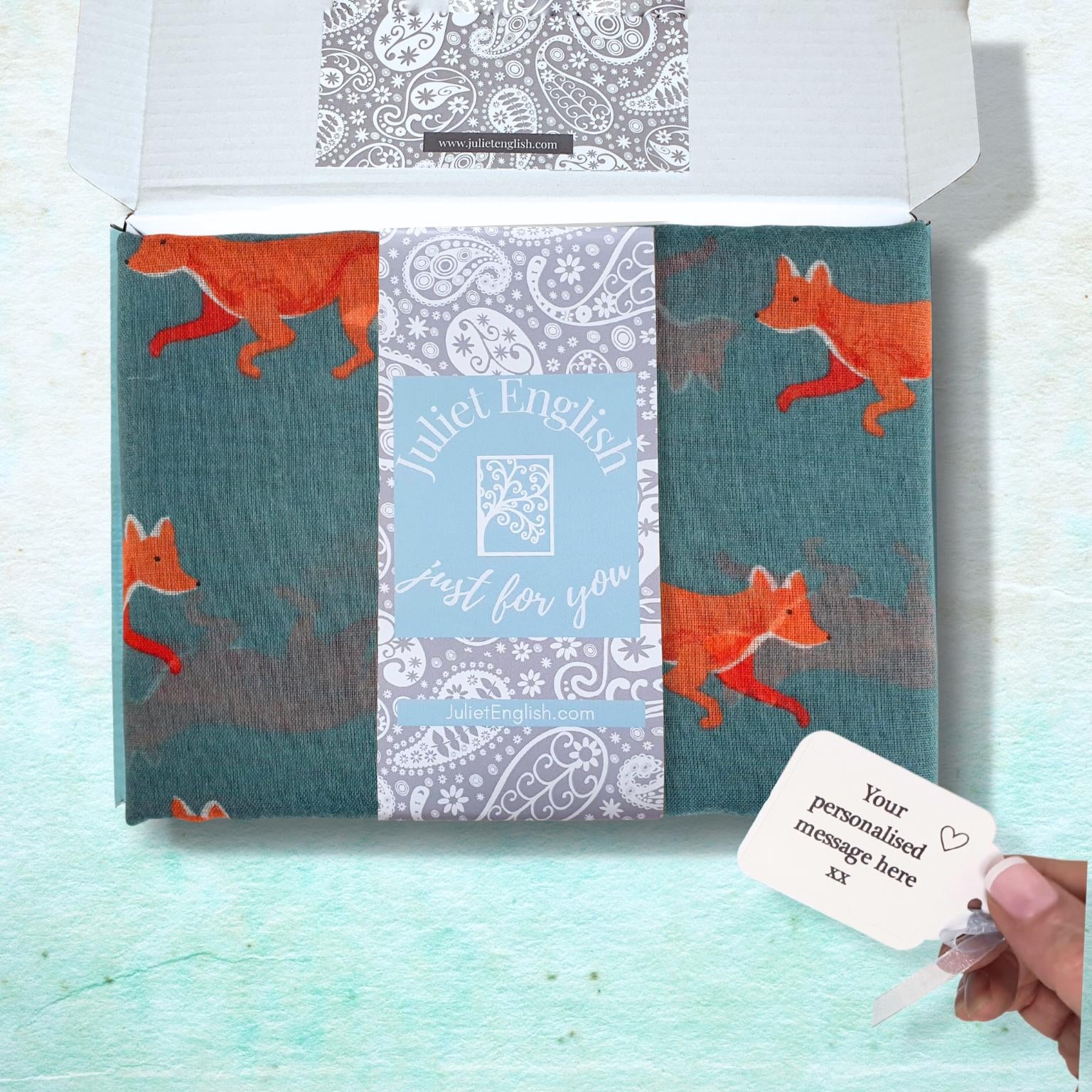 A  box on a light aqua textured background containing a teal green scarf with orange foxes and a central decorative band reading 'Juliet English just for you."