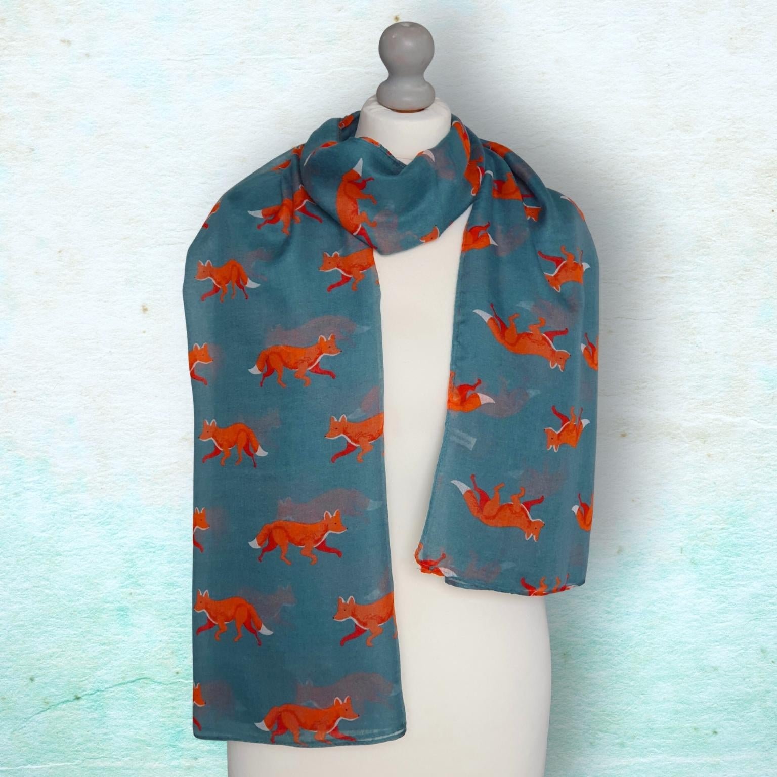 A teal blue scarf with a repeating pattern of orange foxes, displayed on a white mannequin torso against a textured light blue background.