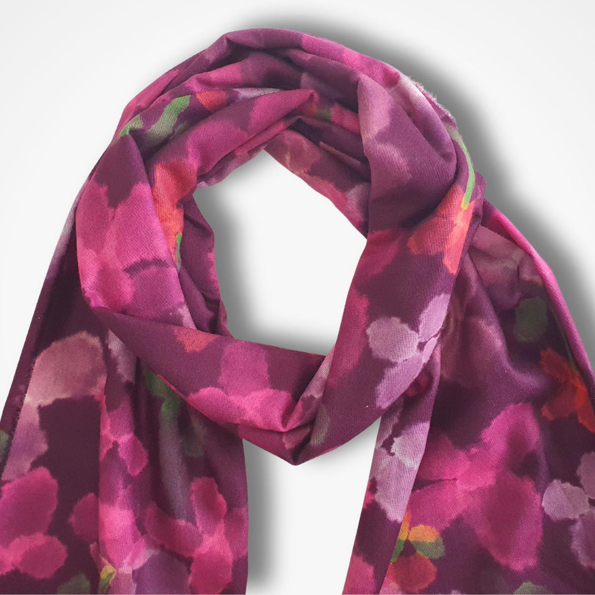 A medium weight scarf with a watercolour-style floral pattern in shades of magenta, pink, and purple, draped against a light gray background.