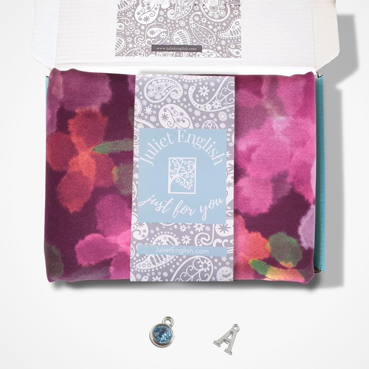 An open gift box containing a folded scarf with a floral print, a light blue central label reading 'Juliet English just for you,' and two small silver charms below on a white background.