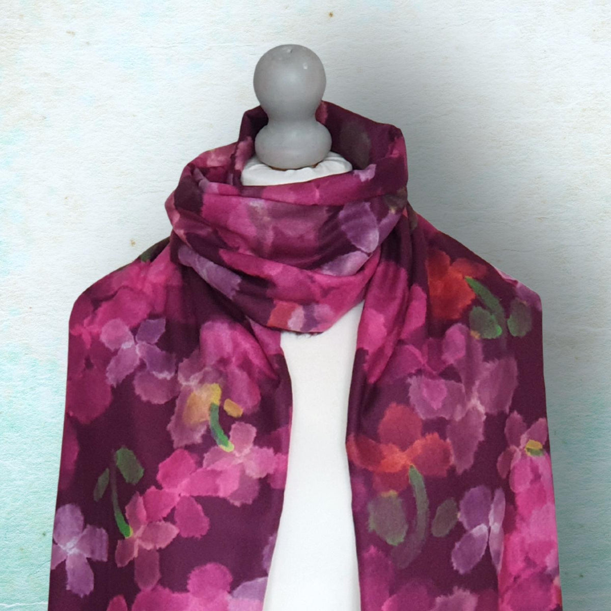 A floral scarf in shades of magenta, pink, and purple with green and orange accents, draped on a white dress form mannequin against a textured light blue background.