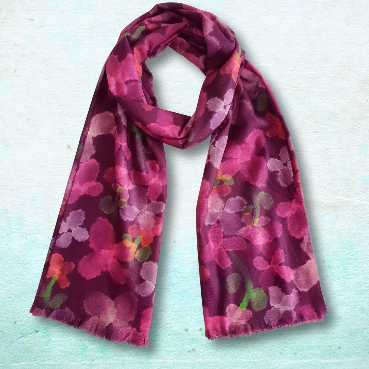 A lightweight scarf with a watercolor-style floral pattern in shades of magenta, pink, and purple, accented with small green and orange details, laid flat on a textured light blue background.