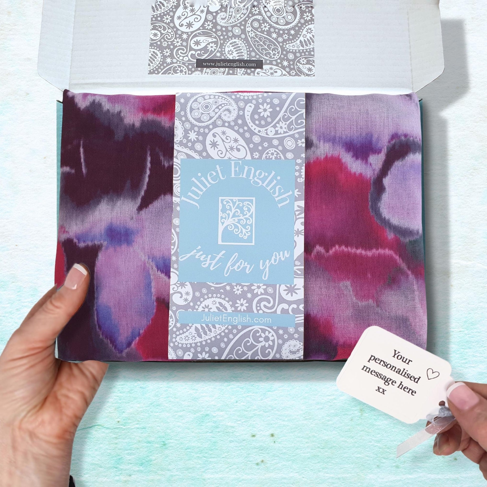 Hand holding a gift box containing a floral scarf with a personalised gift tag on a light blue background